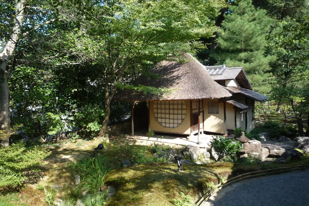 A thatched tea house with a round window among pines, moss and stones in a temple garden